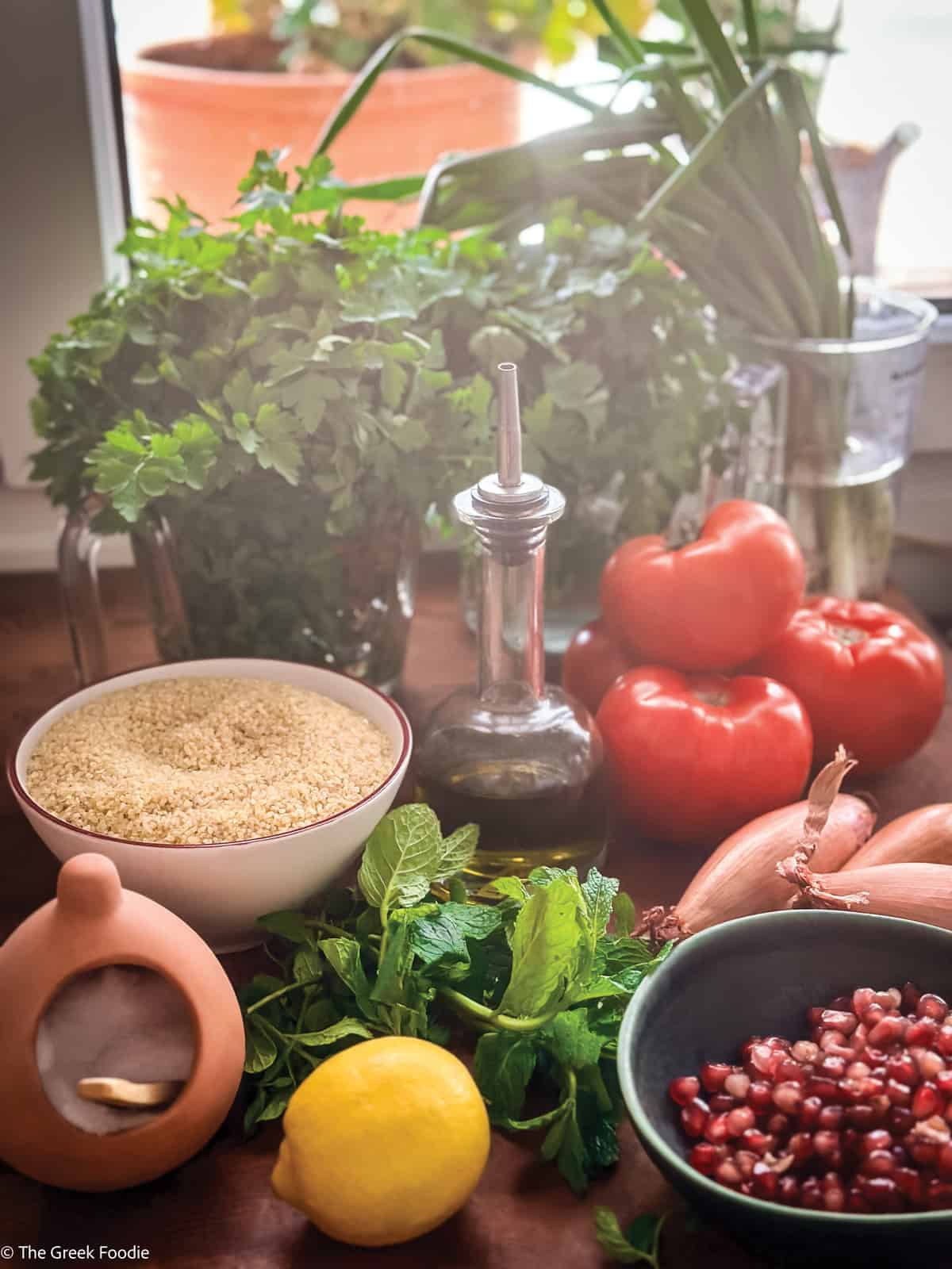 In front of a window on a table are bunches of parsley, a bowl with bulgur wheat, tomatoes, a lemon, pomegranate seeds and mint.