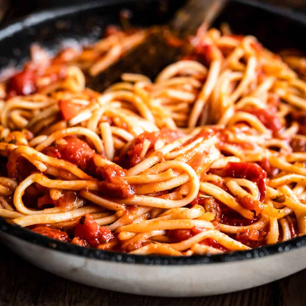 A plate with tomato garlic pasta and a wooden spoon, at the back fresh tomatoes in a bowl.