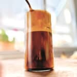A tall glass with frappe coffee in front of an open window.