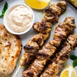 Three pork skewers on a plate with cherry tomatoes, grilled pita , olives, feta and a lemon wedge.