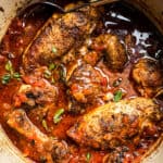 A serving platter with chicken in red sauce, some herbs and two spoons.