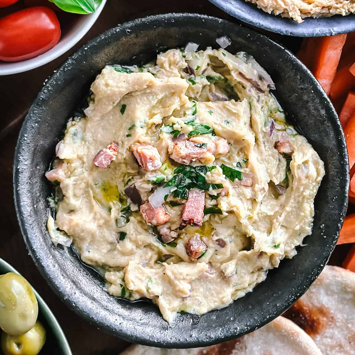 A bowl with hummus with diced charred bacon between vegetables and pitas.