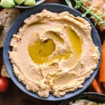 A bowl of hummus, partial view of vegetables and herbs around it.