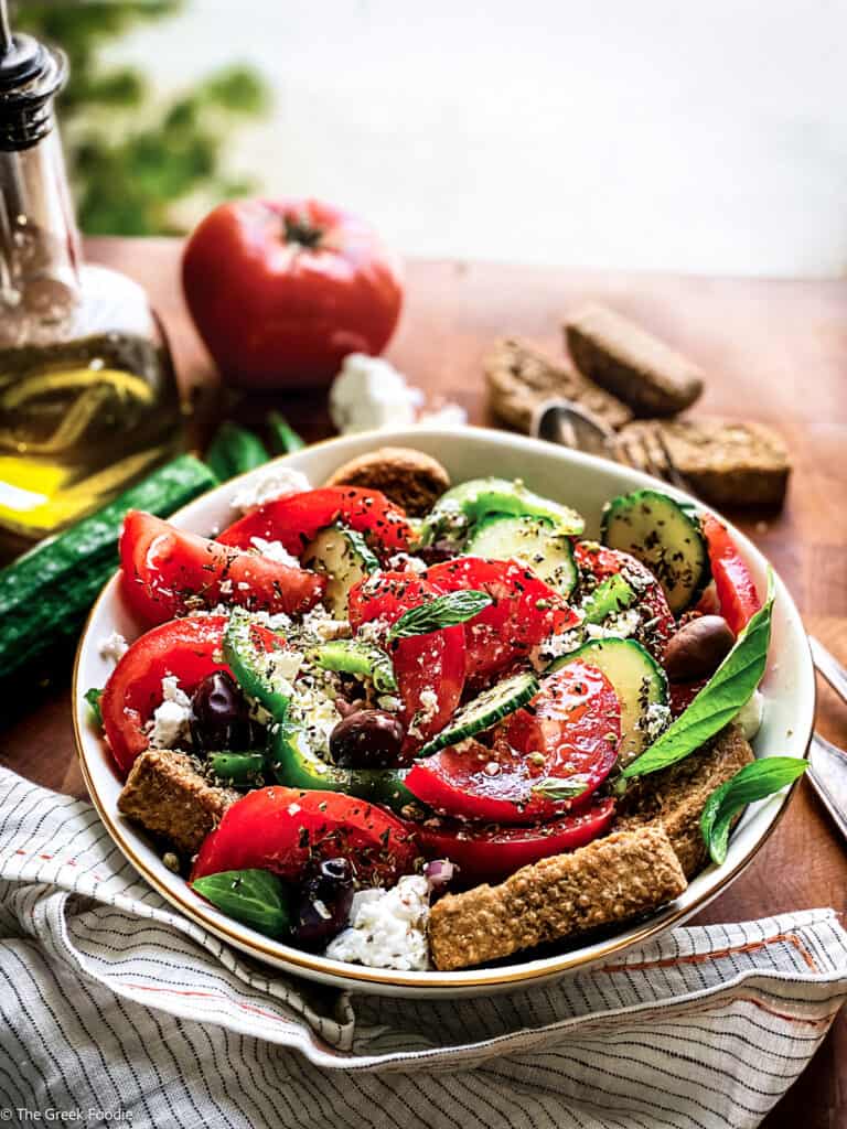 A plate with greek salad, a fork and a spoon an olive oil bottle, feta pieces and rusks, and fresh basil leaves.