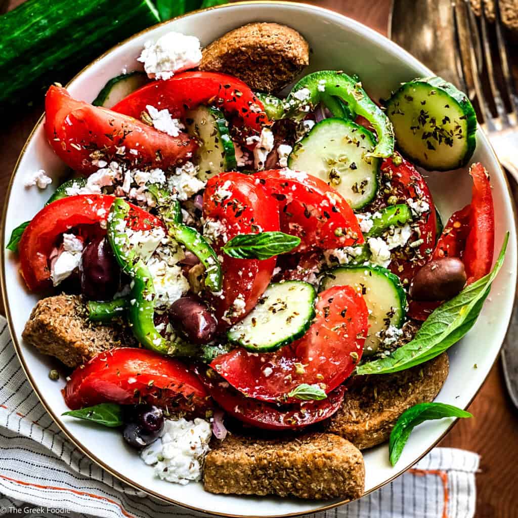 A plate with greek salad, a fork and a spoon an olive oil bottle, feta pieces and rusks, and fresh basil leaves.