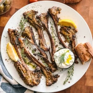 Overhead of grilled Greek lamb chops (paidakia) on a white plate with a lemon wedge, fresh oregano, rosemary, and a bowl of tzatziki