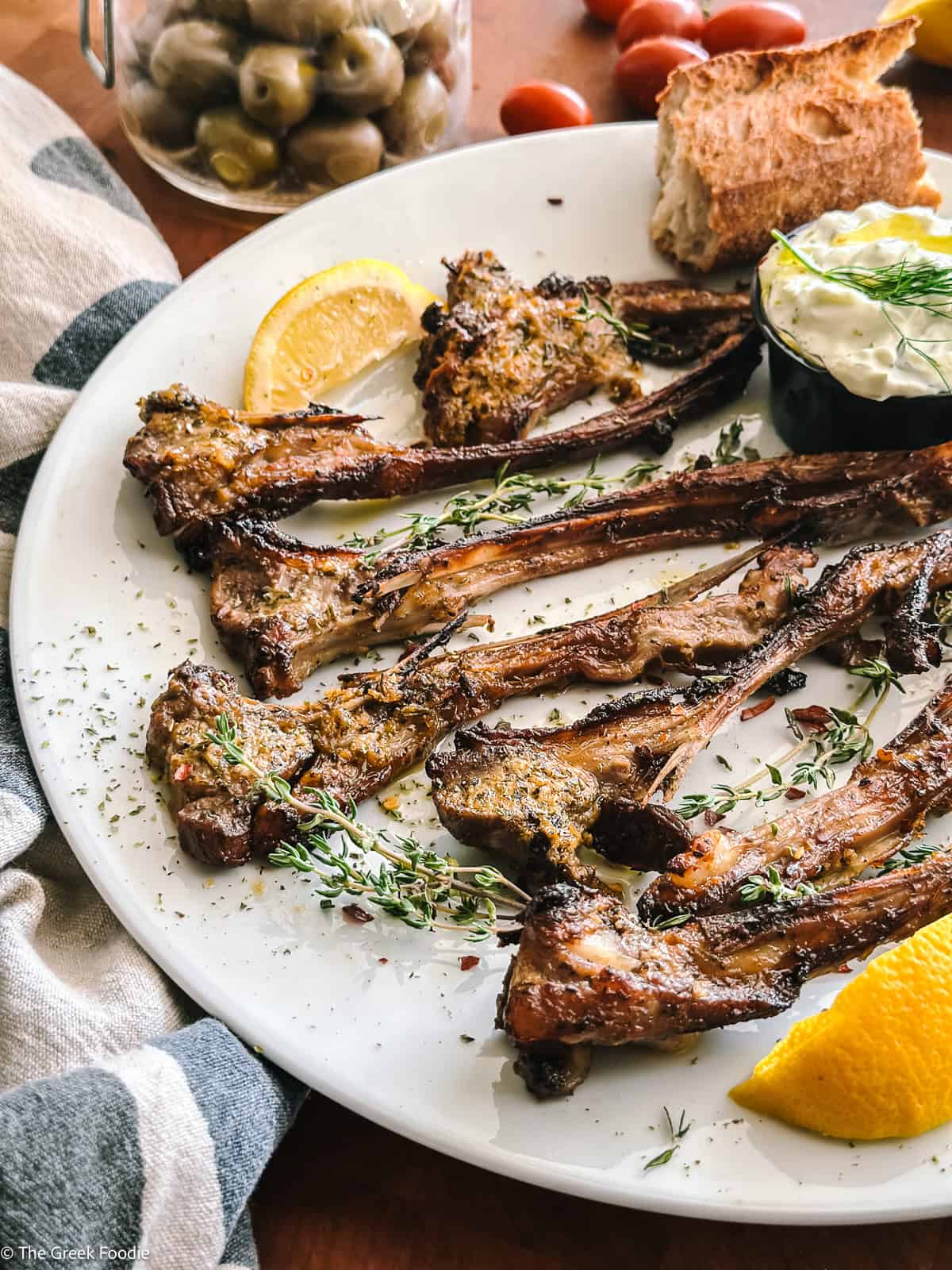 Greek lamb chops with tzatziki sauce, lemon, bread, and fresh herbs on a white plate.