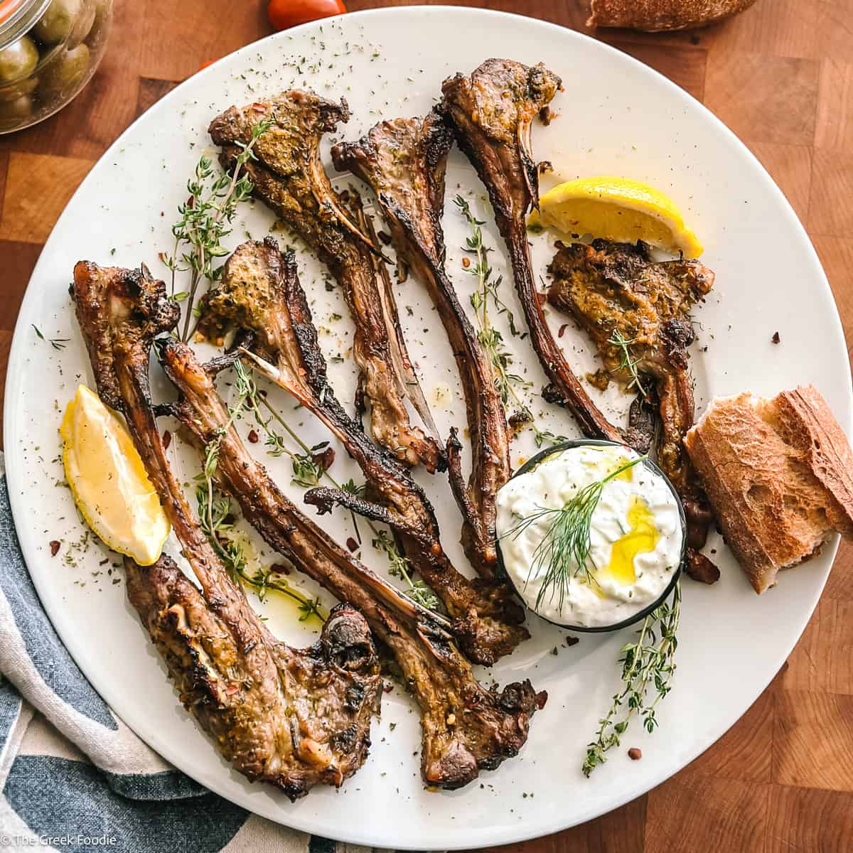 Overhead of grilled Greek lamb chops (paidakia) on a white plate with a lemon wedge, fresh oregano, rosemary, and a bowl of tzatziki