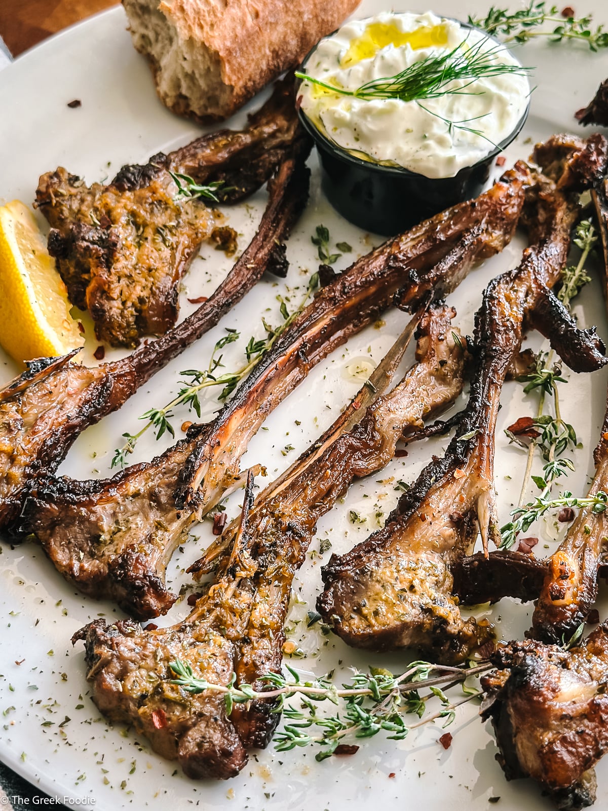 Greek lamb chops with tzatziki sauce, lemon, pita, and fresh herbs on a white plate.