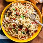 A plate with garlic chili tuna pasta and utensils. On the table lemon, bread and a cloth napkin.