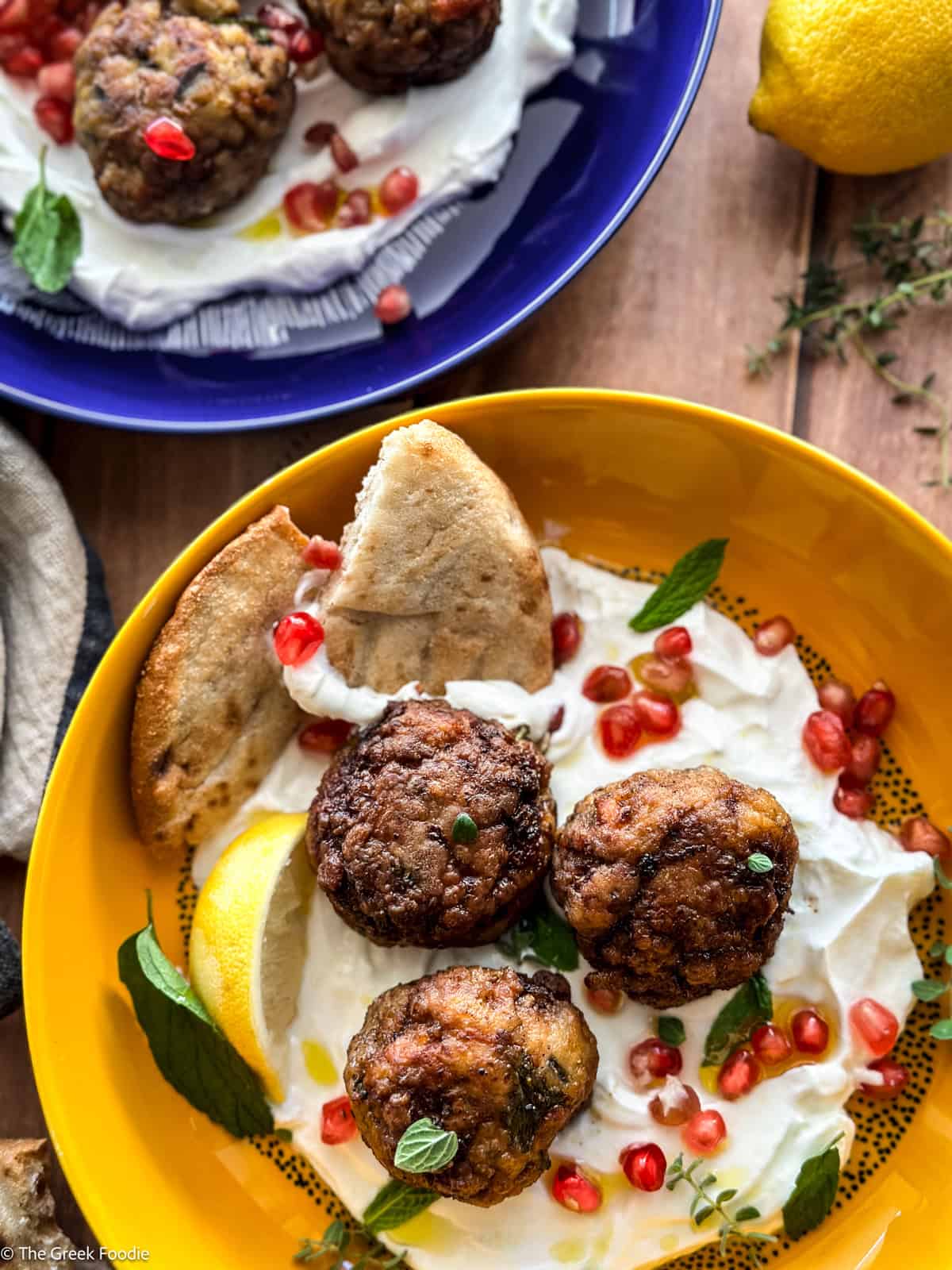 Three lamb meatballs on yogurt, garnished with pomegranate seeds, herbs and pita pieces in a yellow plate. At the back another plate with meatballs.