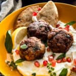 Three lamb meatballs on yogurt, garnished with pomegranate seeds, herbs and pita pieces in a yellow plate. At the back another plate with meatballs.