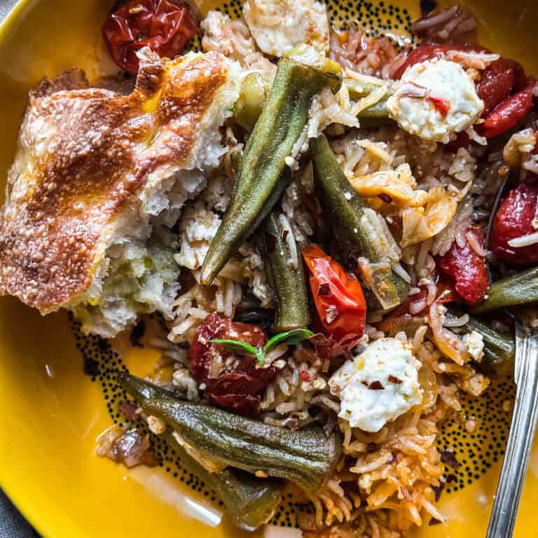 Baked rice with okra and feta, with a piece of bread on a plate with a fork, on a table with more bread and a cloth napkin.