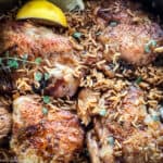 A skillet with chicken and rice, lemon wedges and herbs on a table with a cloth napkin.