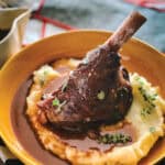 A Greek lamb shank over mashed potatoes in a plate, on a table with a cloth napkin.