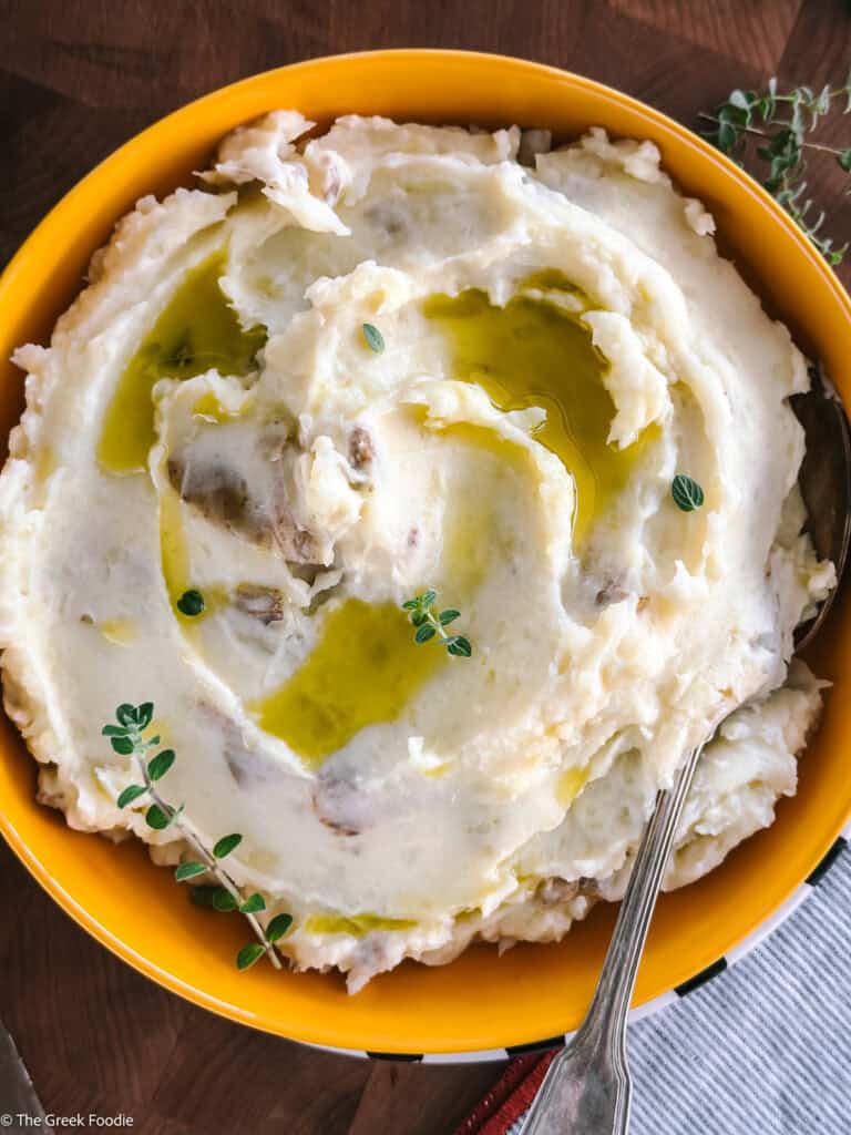 A yellow bowl with olive oil mashed potatoes, herbs and a spoon.