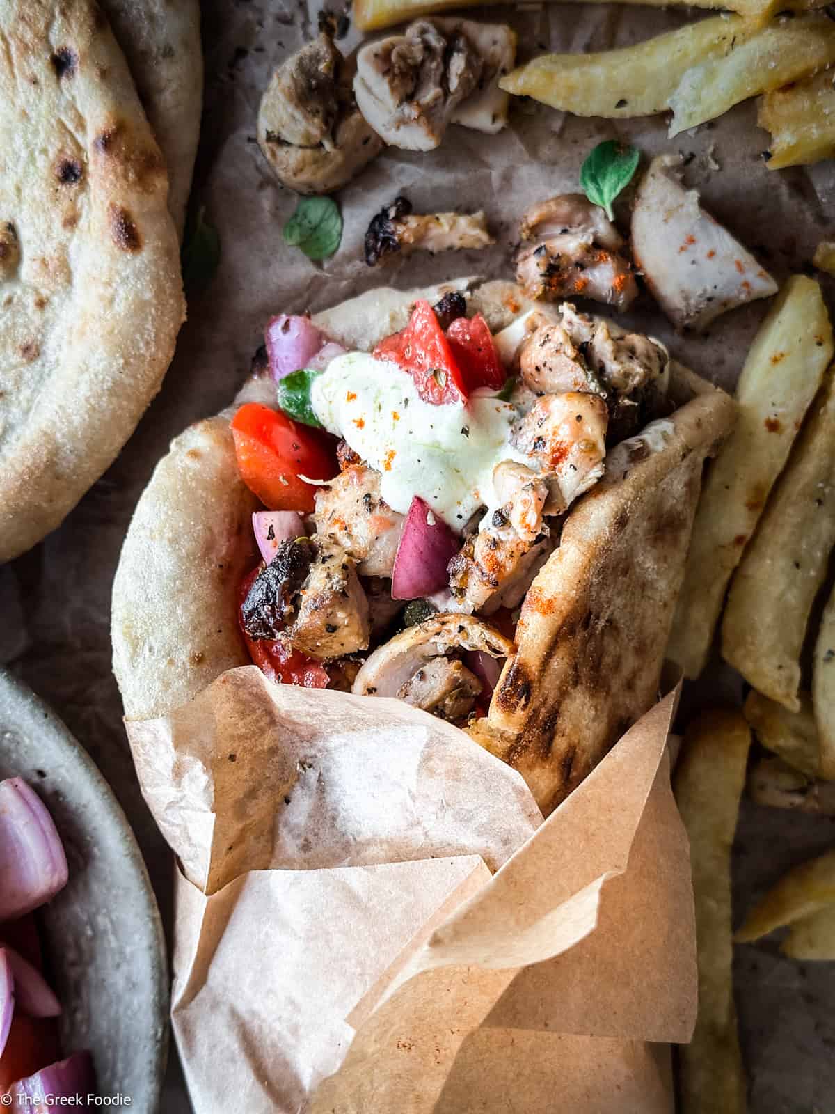 A chicken gyros souvlaki wrapped on paper, next to pita and fries.