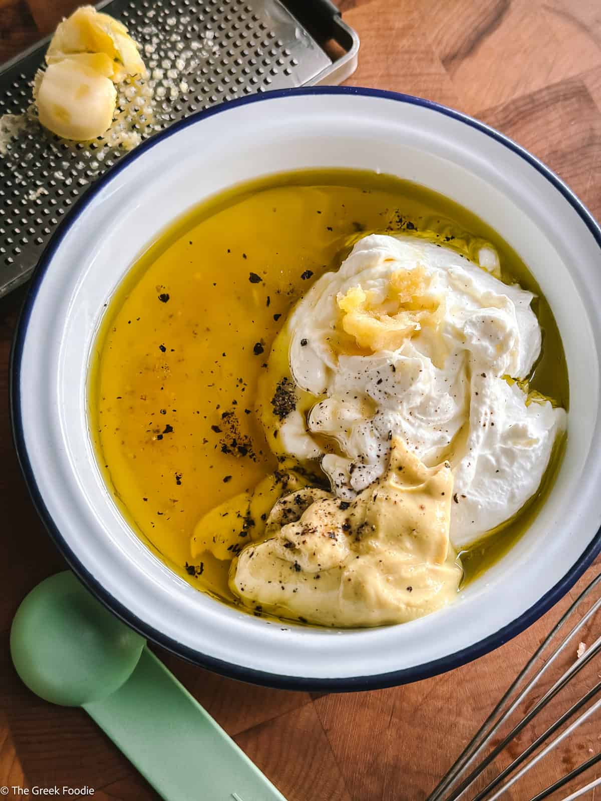 Olive oil, yogurt, mustard and seasonings in a shallow bowl with kitchen utensils around on a wooden table.