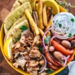 A gyro bowl with pita, chicken gyro, sliced tomatoes, tzatziki, olives and fries on a table.