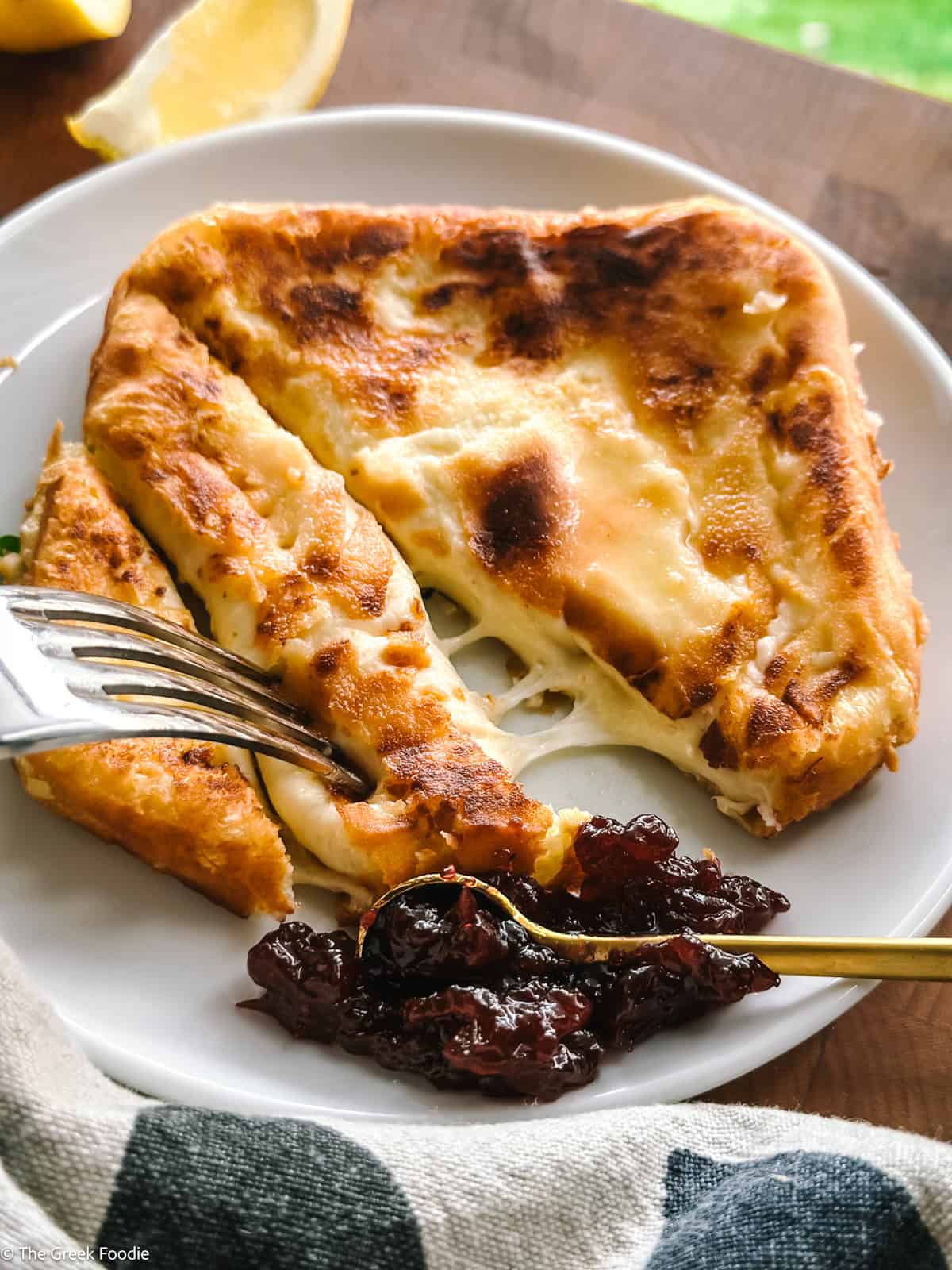 Saganaki Greek fried cheese sliced with a fork and a spoon with chutney on a plate, a lemon wedge and a cloth napkin on a table.