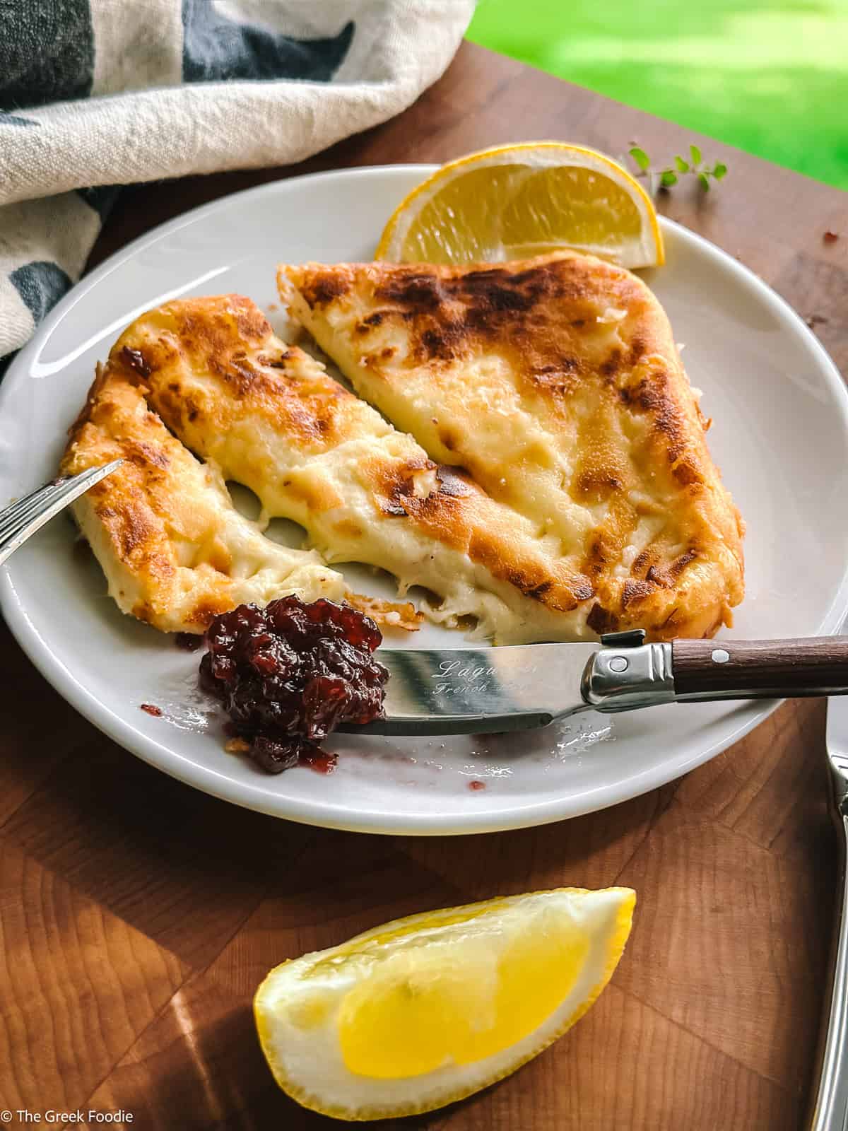 Saganaki Greek fried cheese sliced with a fork and a spoon with chutney on a plate, a lemon wedge and a cloth napkin on a table.