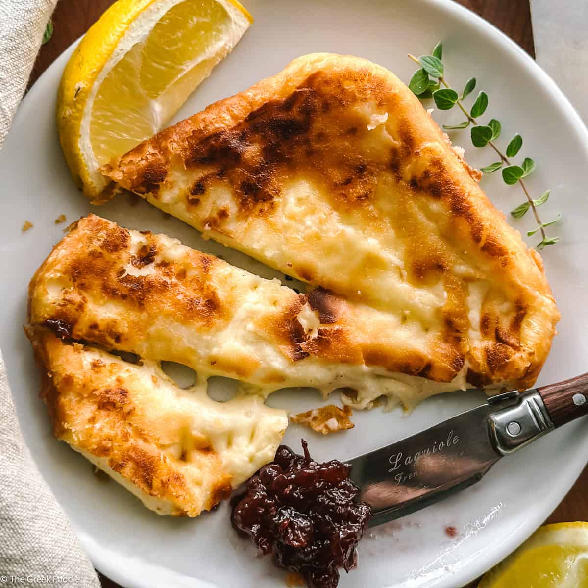 Saganaki Greek fried cheese sliced with a fork and a spoon with chutney on a plate, a lemon wedge and a cloth napkin on a table.