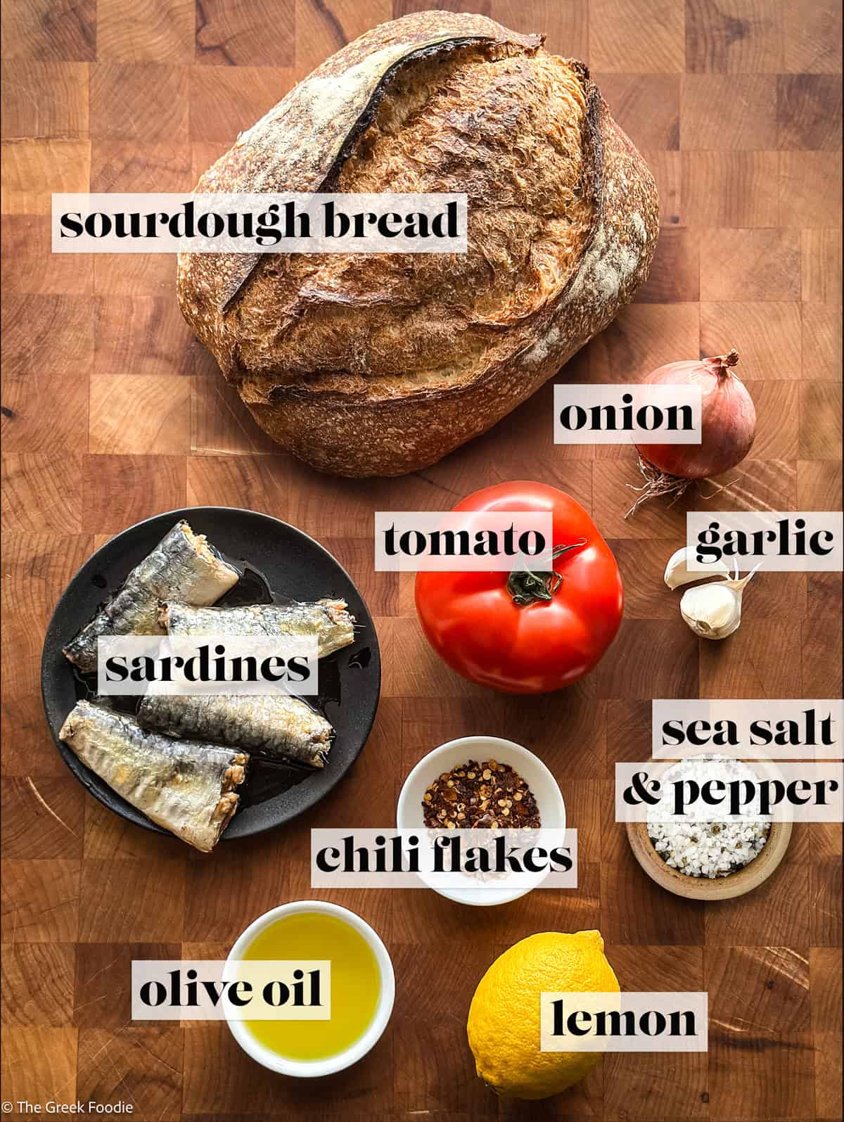 Sourdough bread, sardines, olive oil, sea salt and pepper, a lemon, a tomato and an onion on a wooden surface.
