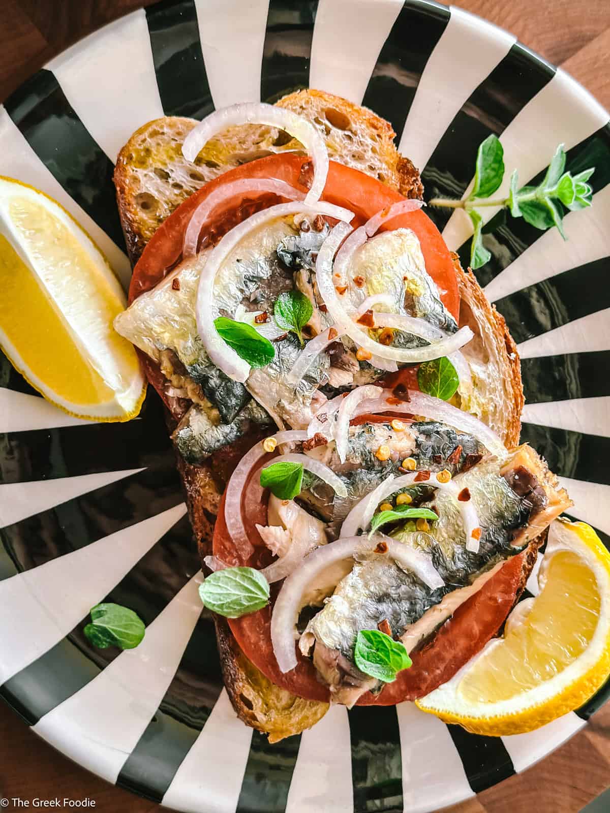 Sardines on toast and lemon wedges on a black and white plate.