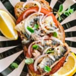 Sardines on toasted sourdough bread and lemon wedges on a black and white plate.
