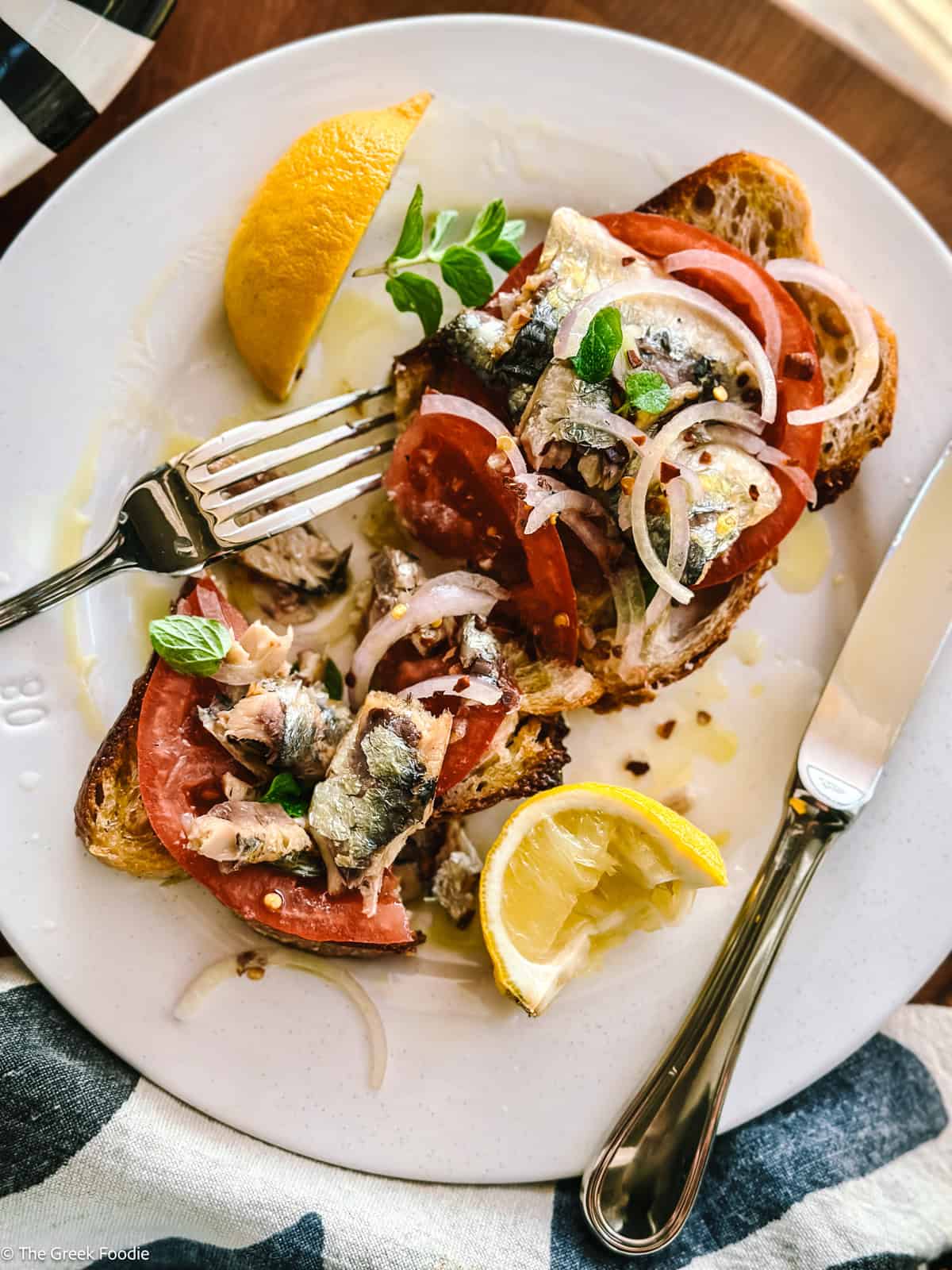 Sardines on toast cut on a plate with a fork and a knife and lemon wedges.