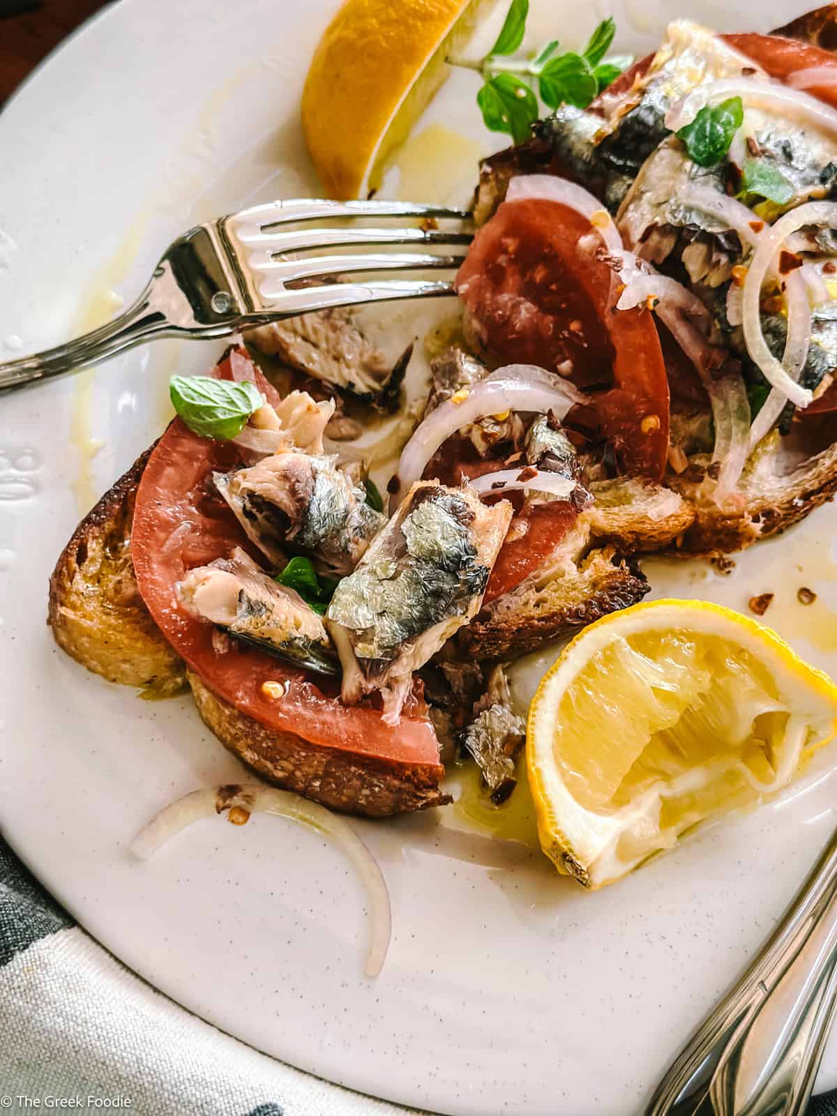 Sardines on toast cut on a plate with a fork and a knife and lemon wedges.
