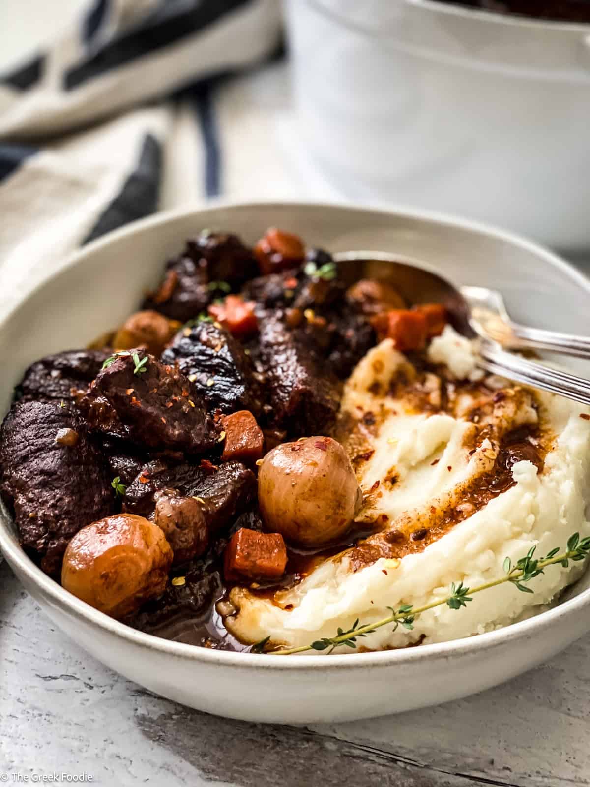 A plate with red wine beef stew and mashed potatoes. At the back a white pot and a cloth napkin.