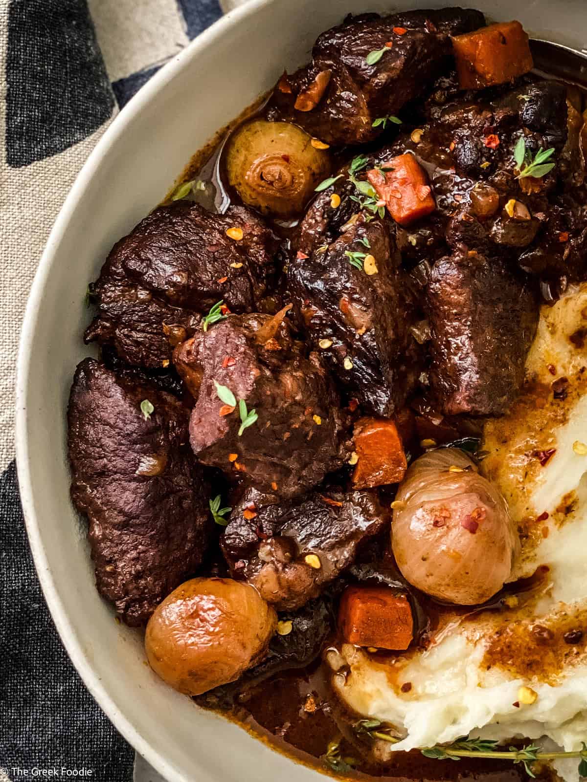 A plate with red wine beef stew and mashed potatoes. At the back a white pot and a cloth napkin.