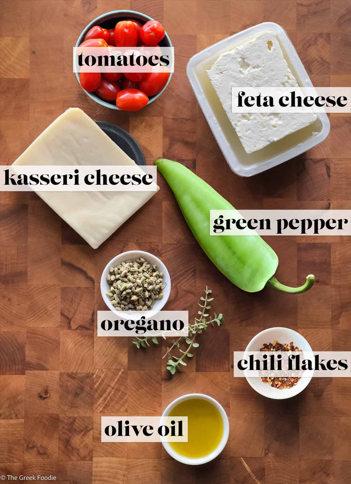 Feta cheese, tomatoes, Kasseri cheese, olive oil, chili flakes, oregano and a green pepper on a wooden board.