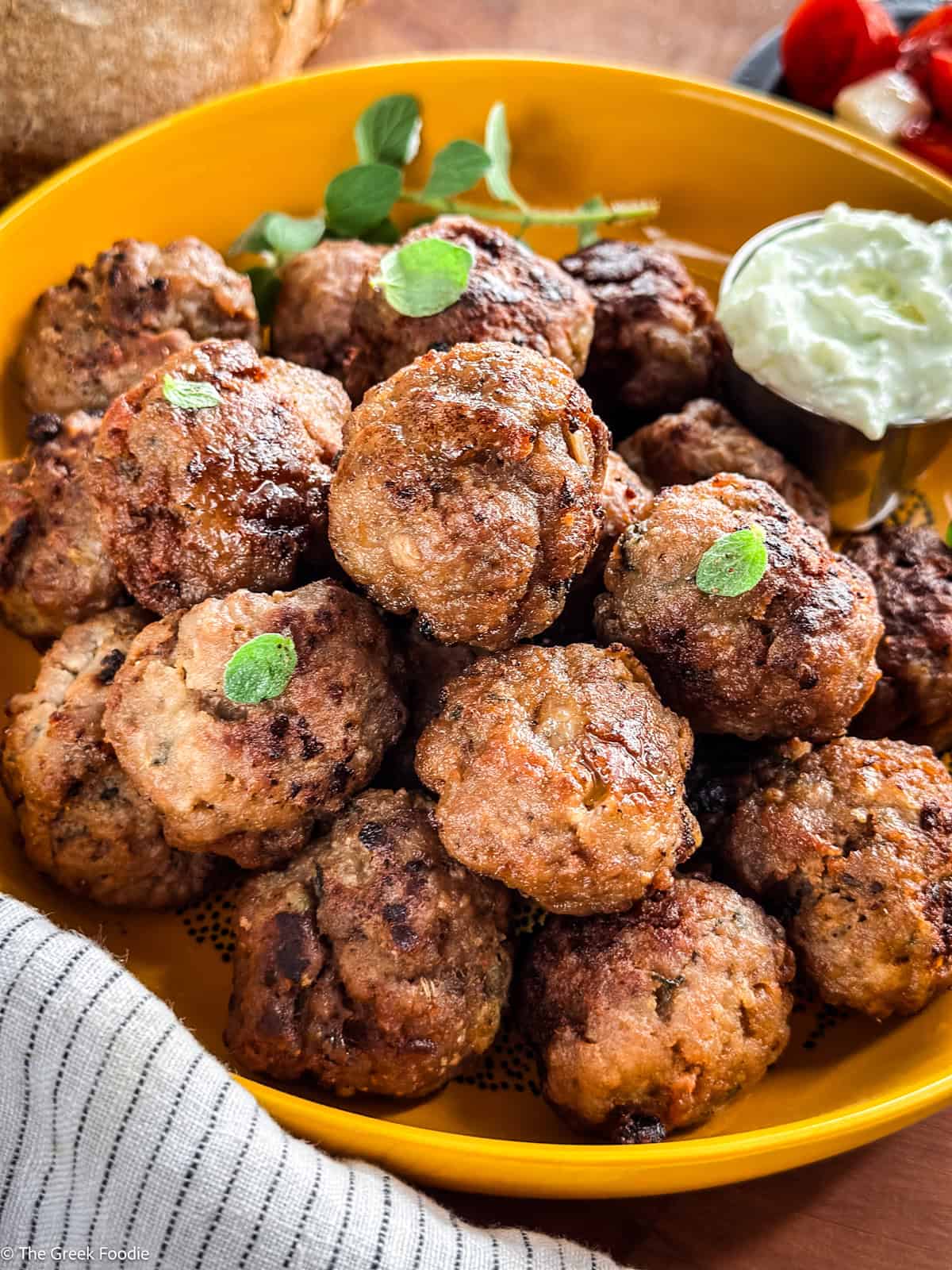 A yellow plate full of meatballs and a small bowl of tzatziki sauce on a table, and a cloth napkin.
