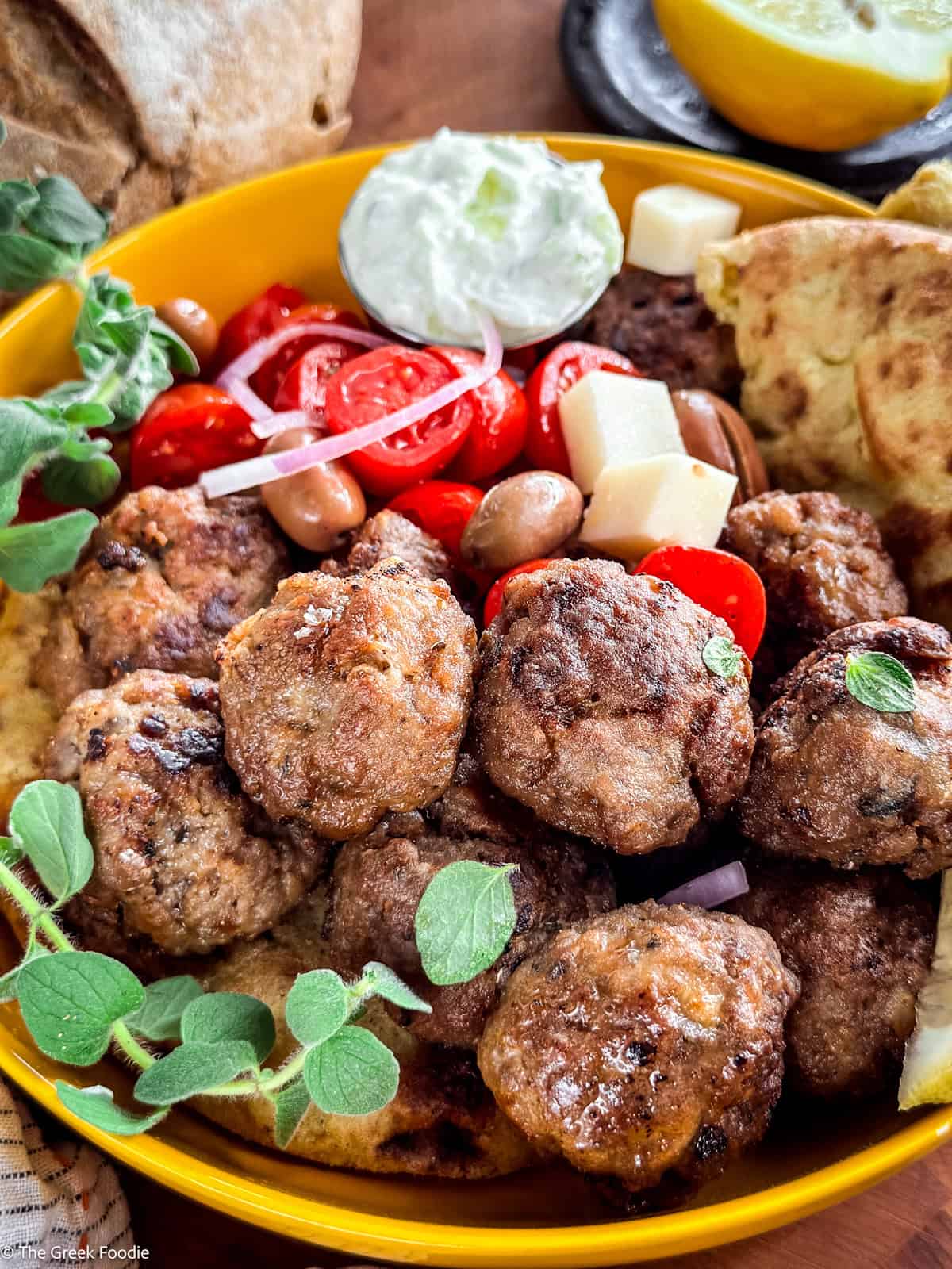 A yellow plate full of meatballs, cherry tomatoes, cubed cheese, herbs and pita on a table.