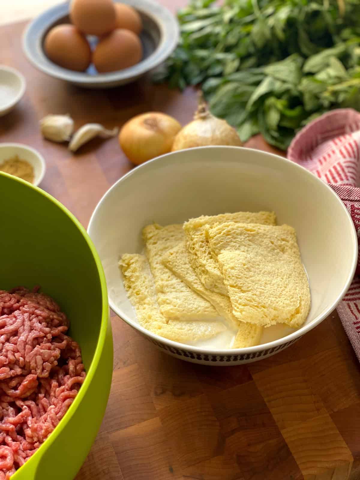 A bowl with milk and pieces of bread on a working surface surrounded with other meatball ingredients.