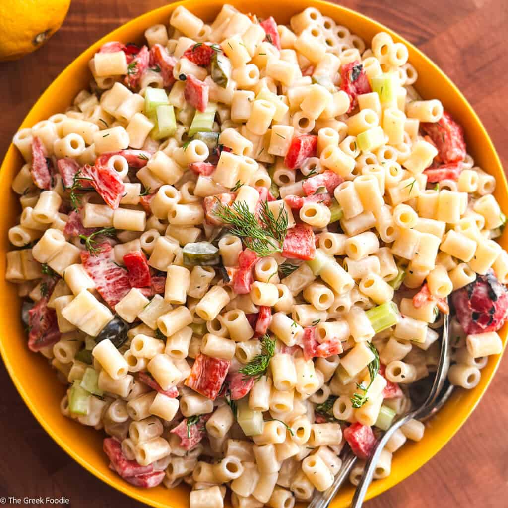 Dill pickle pasta salad with roasted red peppers, celery and fresh dill in a yellow bowl with two serving spoons.