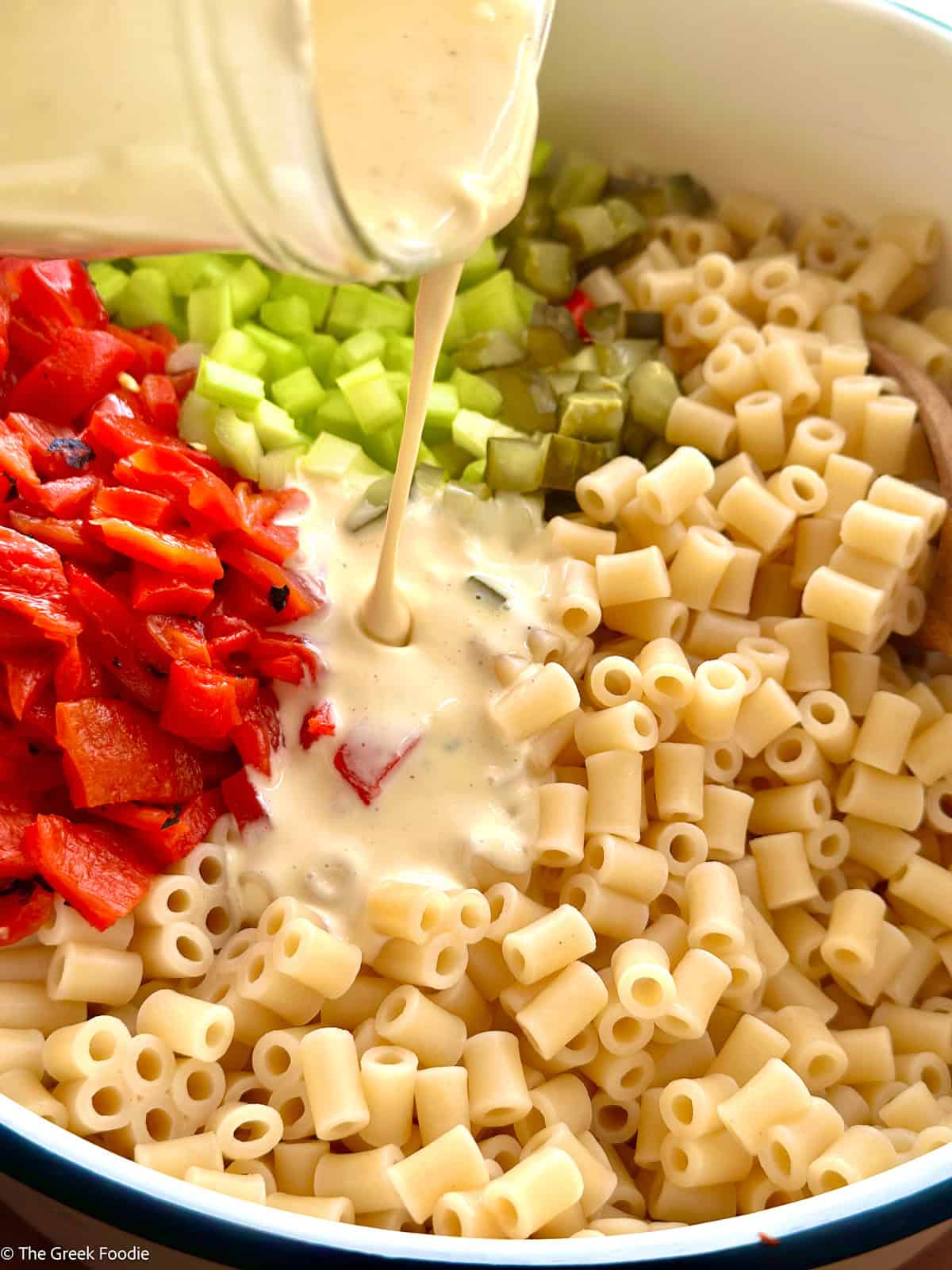Creamy Dijon-lemon mayo dressing being poured over ditalini, diced roasted red peppers, celery and dill pickles in a bowl.