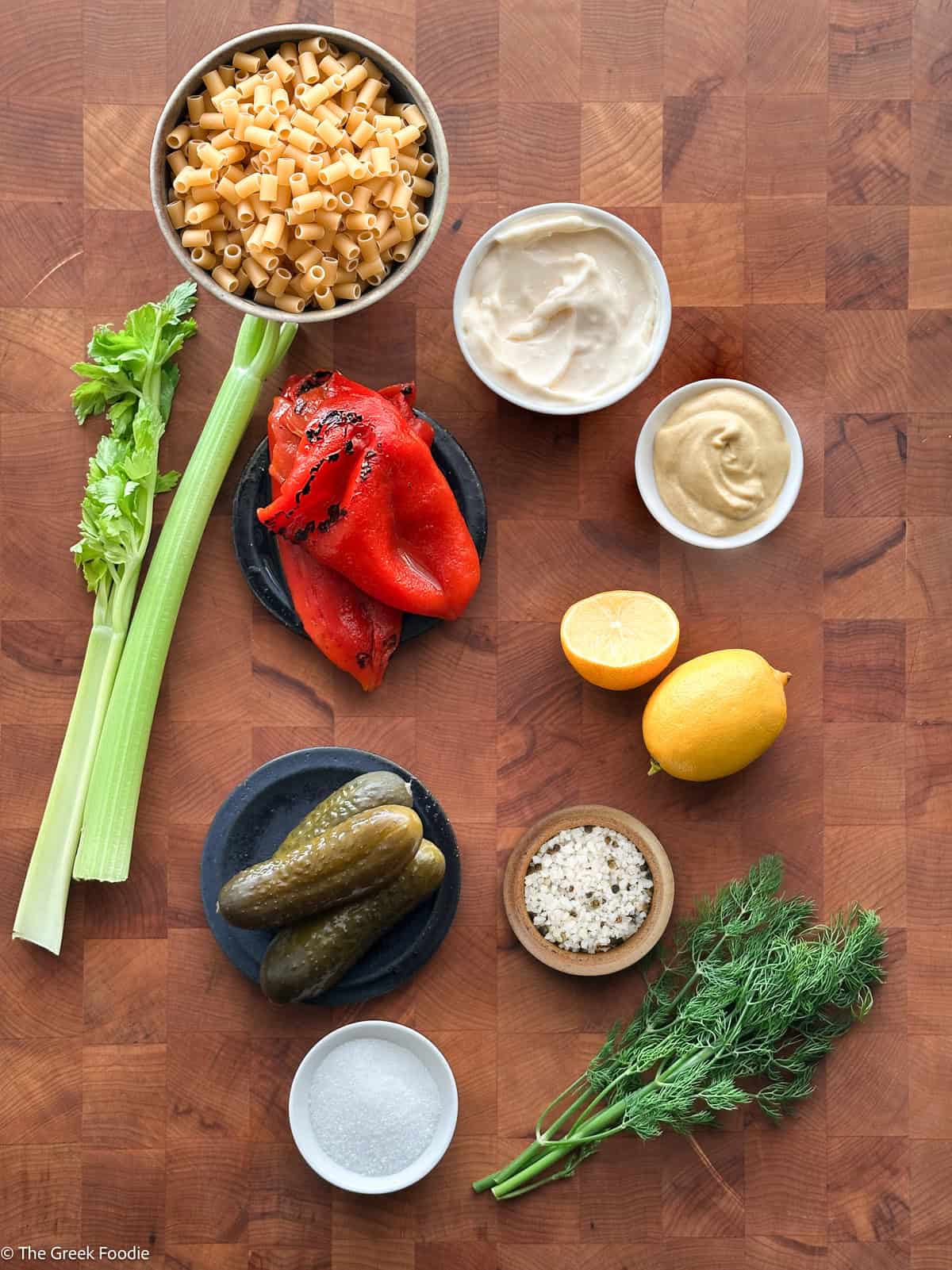 Ingredients for dill pickle pasta salad on a wooden board: ditalini, mayonnaise, Dijon mustard, roasted red peppers, celery, dill pickles, lemons, fresh dill, salt and sugar.