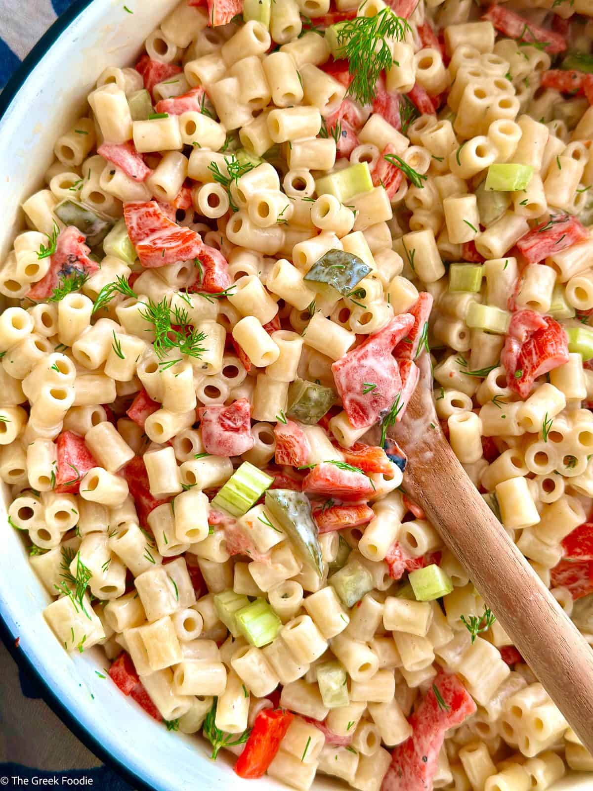 Dill pickle macaroni salad being tossed with a wooden spoon in a white enamel bowl.
