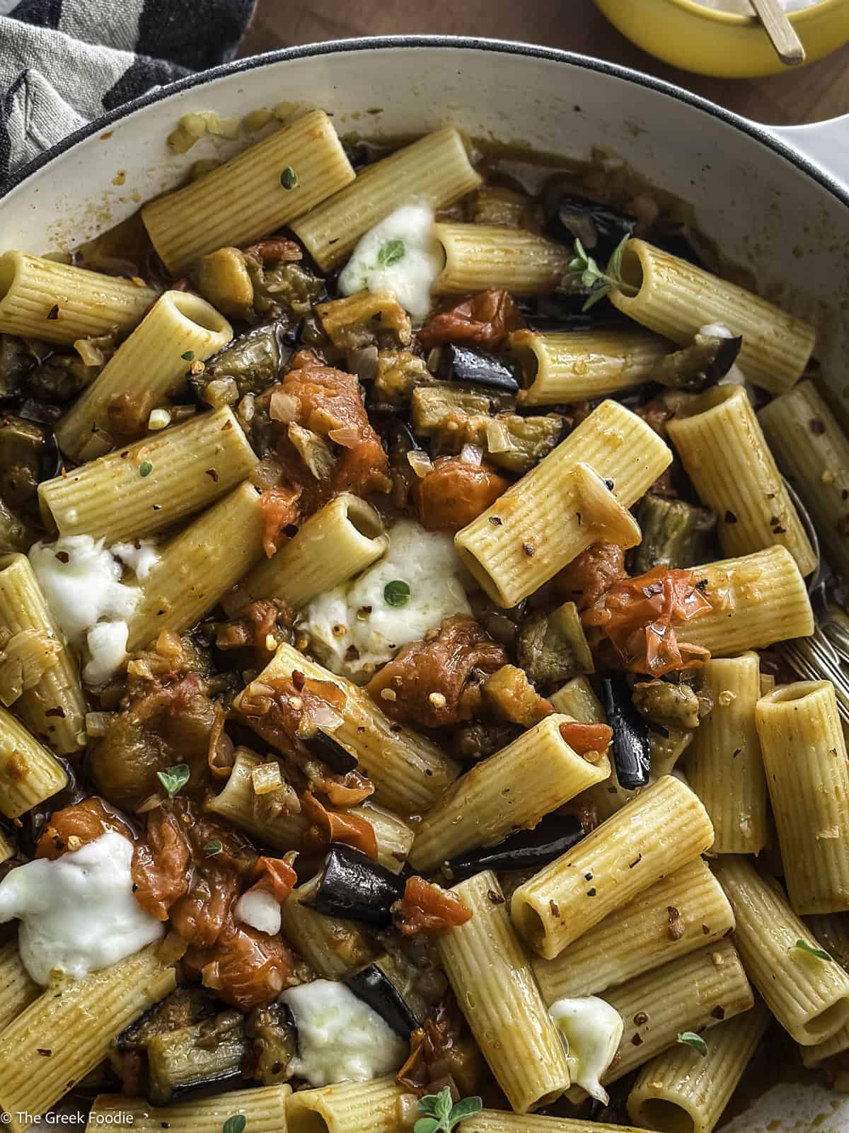 A white pot with eggplant pasta, on a table with a wooden spoon.