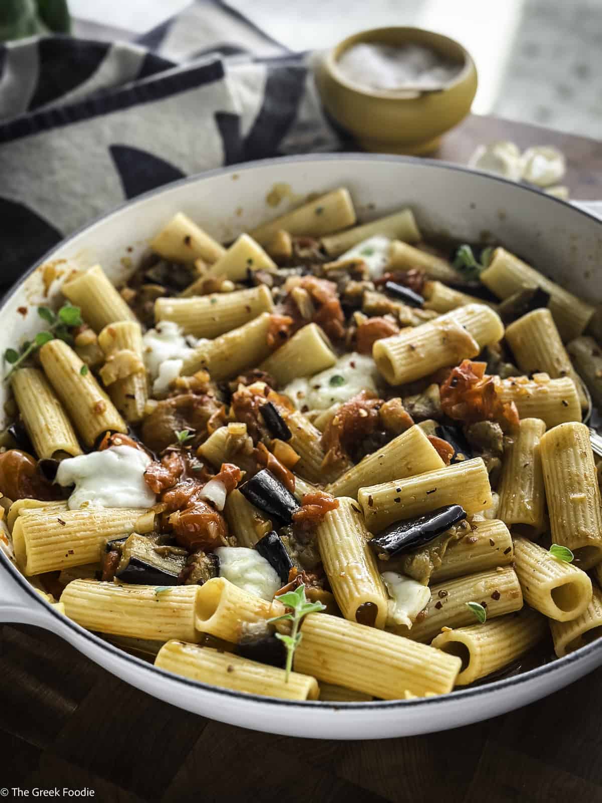 A white pot with eggplant pasta on a table with a cloth napkin and a salt cellar.