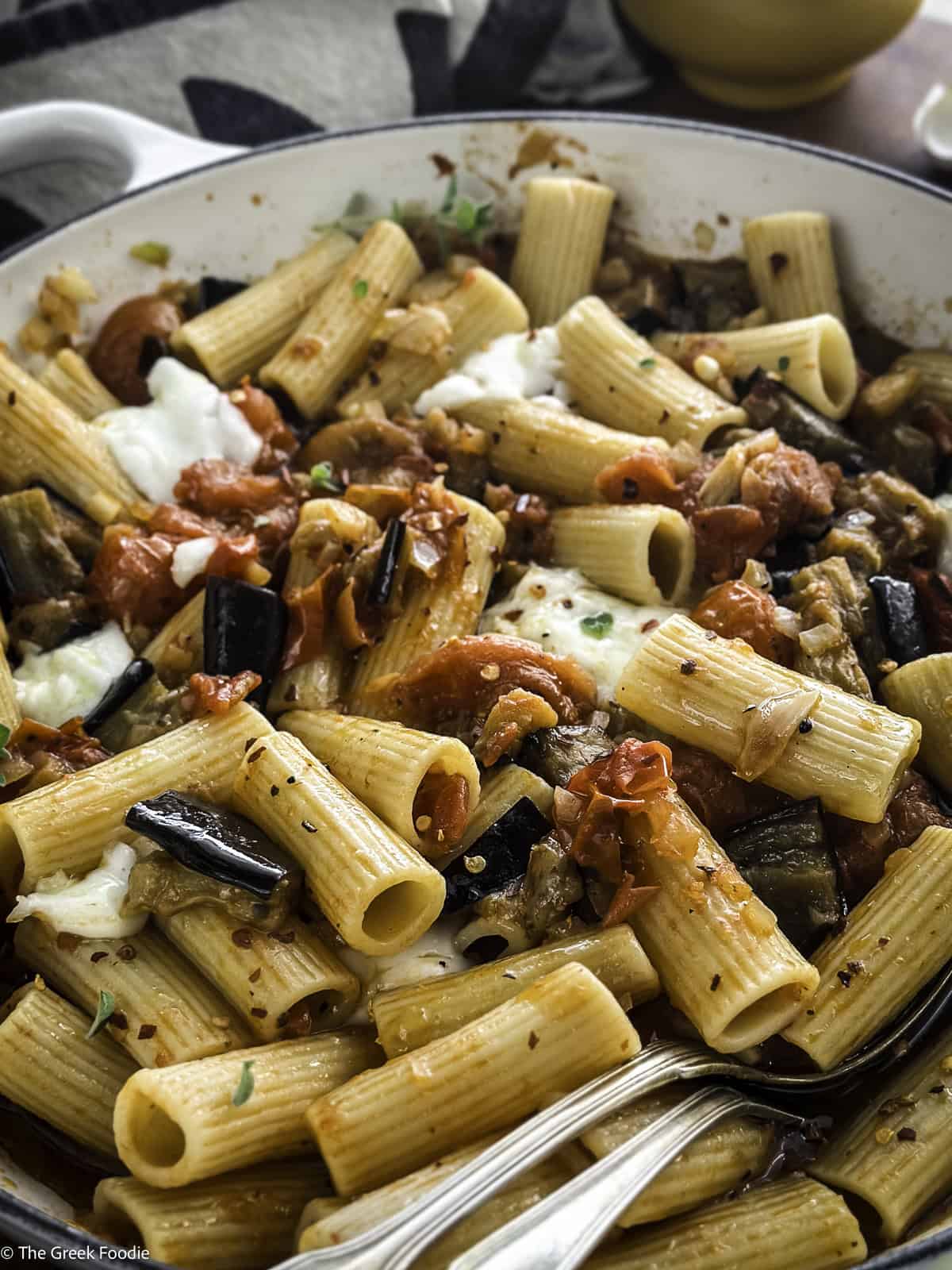 A white pot with eggplant pasta on a table with a cloth napkin and a salt cellar.