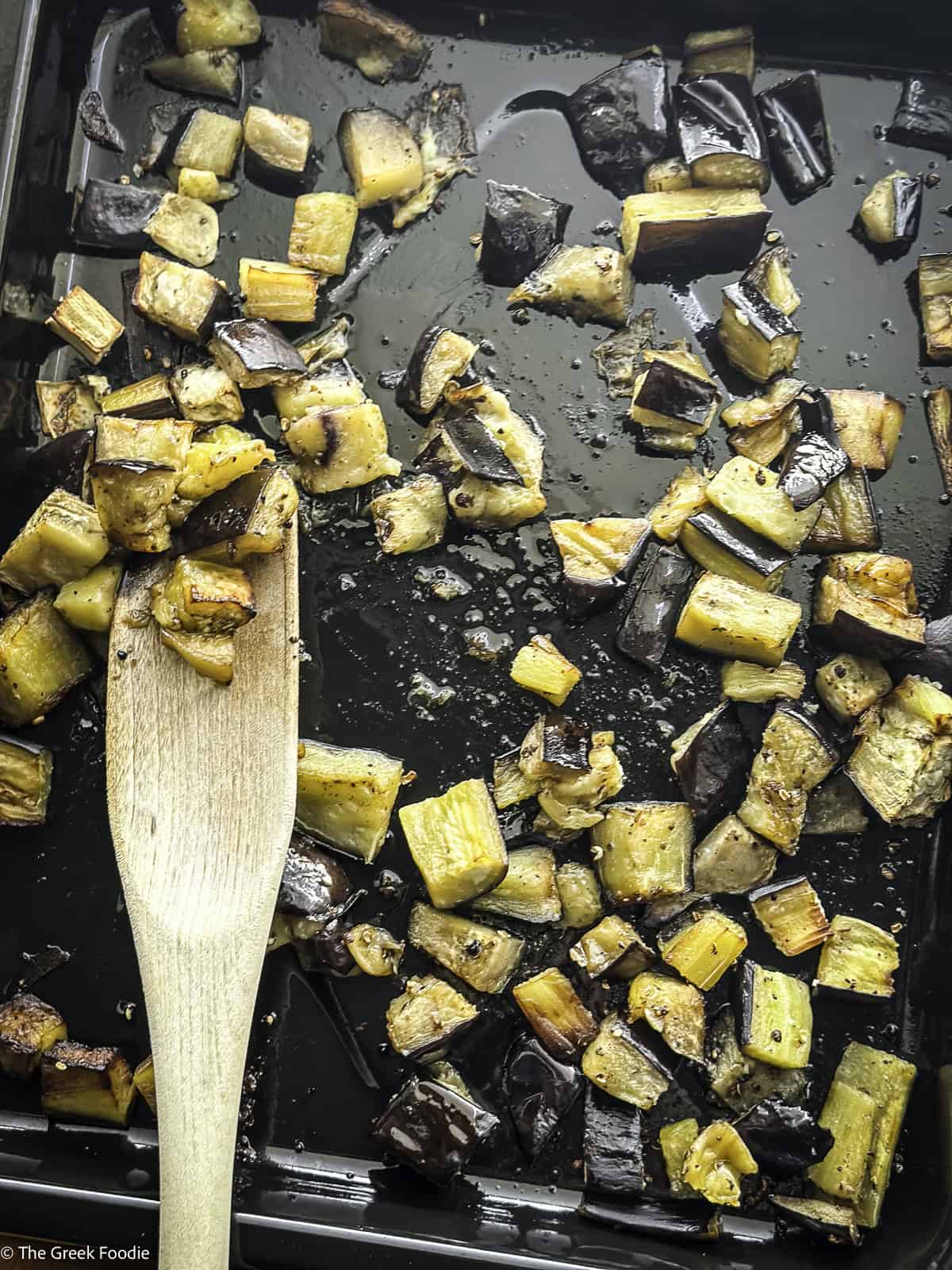 Roasted eggplant cubes dressed with olive oil and seasonings in a roasting pan and a wooden spoon.