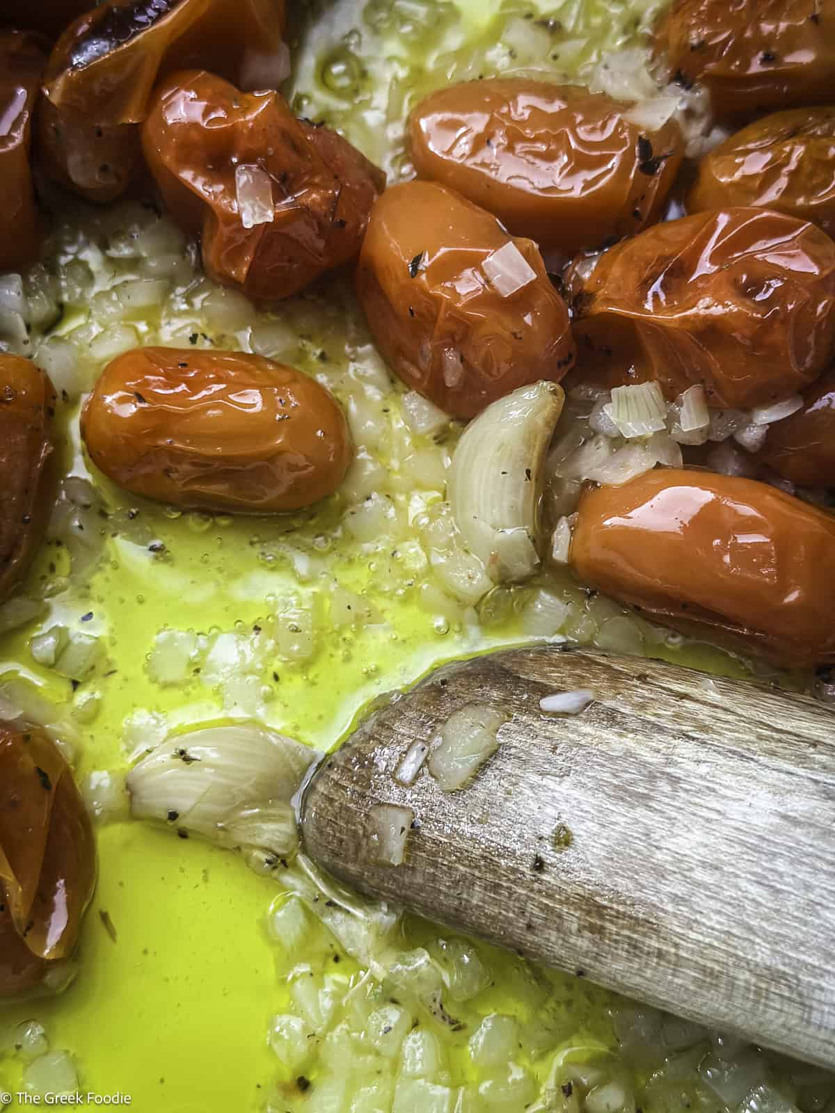 A spoon breaking a garlic clove in a roasting pan with roasted cherry tomatoes.