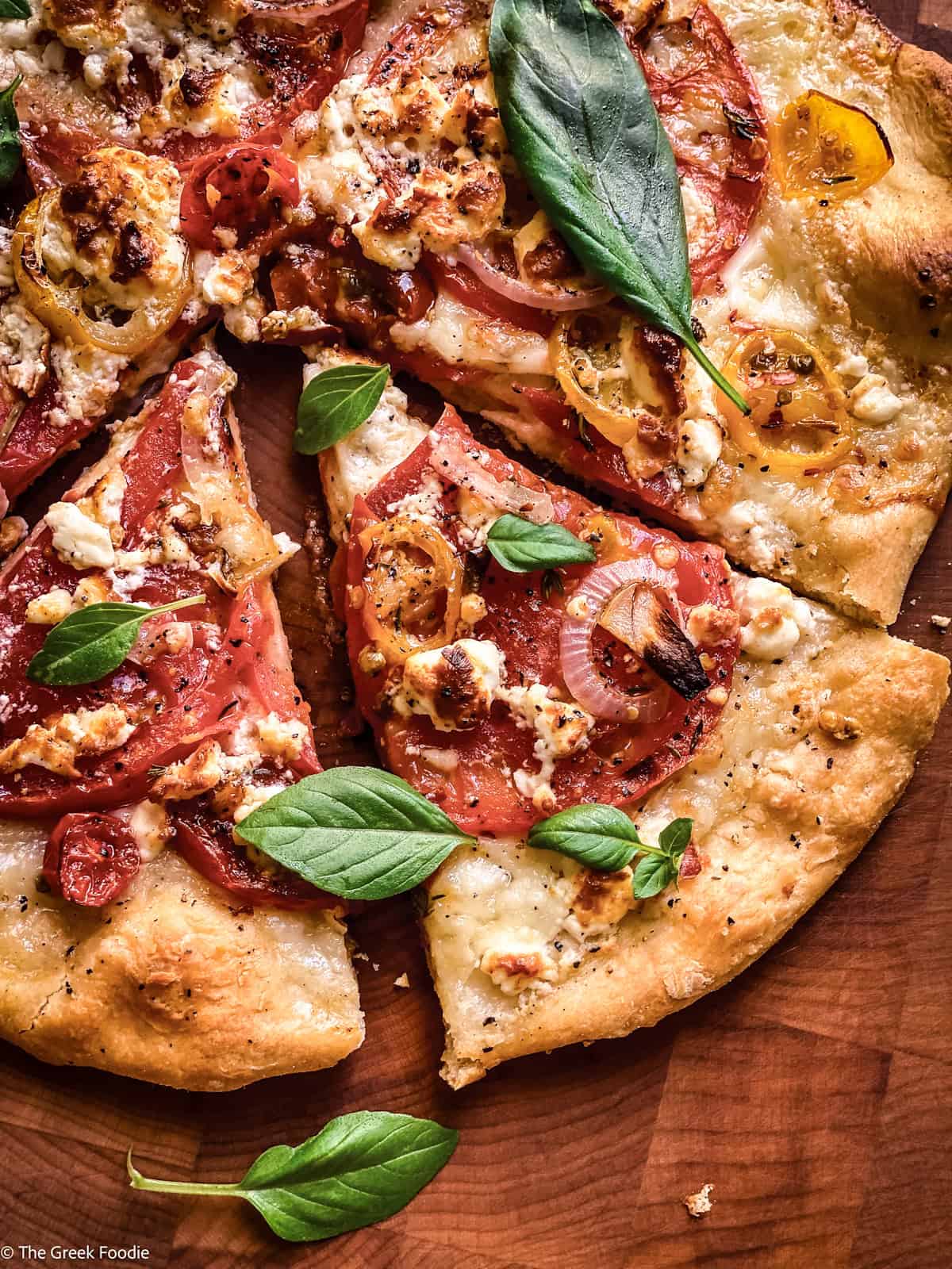 A Greek pizza on a cutting board, two pieces cut and basil leaves scattered on top.