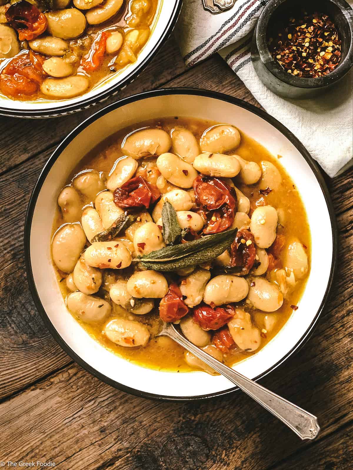 Two plates with Greek gigantes beans, roasted tomatoes and sage leaves and some spoons.