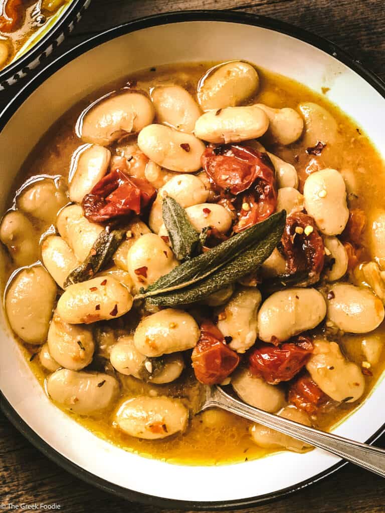 Gigantes beans with roasted tomatoes, two sage leaves in a bowl and a spoon.