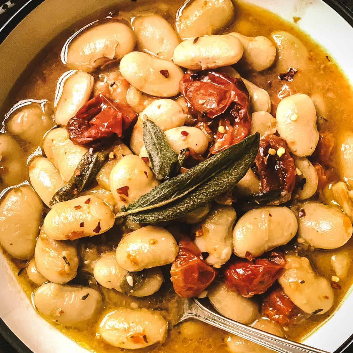Gigantes beans with roasted tomatoes, two sage leaves in a bowl and a spoon.